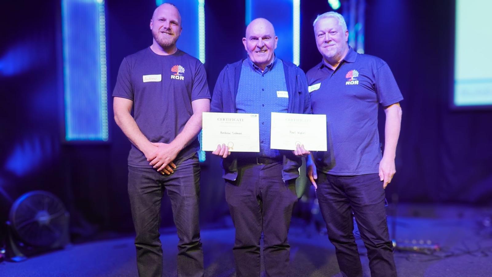 Joel, Andrew Sedman and Paul Wykes receiving award certificates on stage